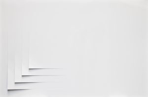 Abstract artwork featuring a series of intricately cut, overlapping white paper shapes arranged in a circular, spiral-like pattern. The layered elements create depth and shadow, adding dimension to the otherwise minimal composition on a white background. The piece invites viewers to explore subtle shifts in light and form, with a delicate interplay of curves and sharp angles.